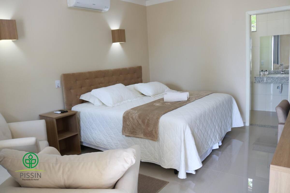Ein Hotelzimmer mit einem großen Bett, einem Nachttisch, einem Sessel und einem durch eine offene Tür einsehbaren Badezimmer.