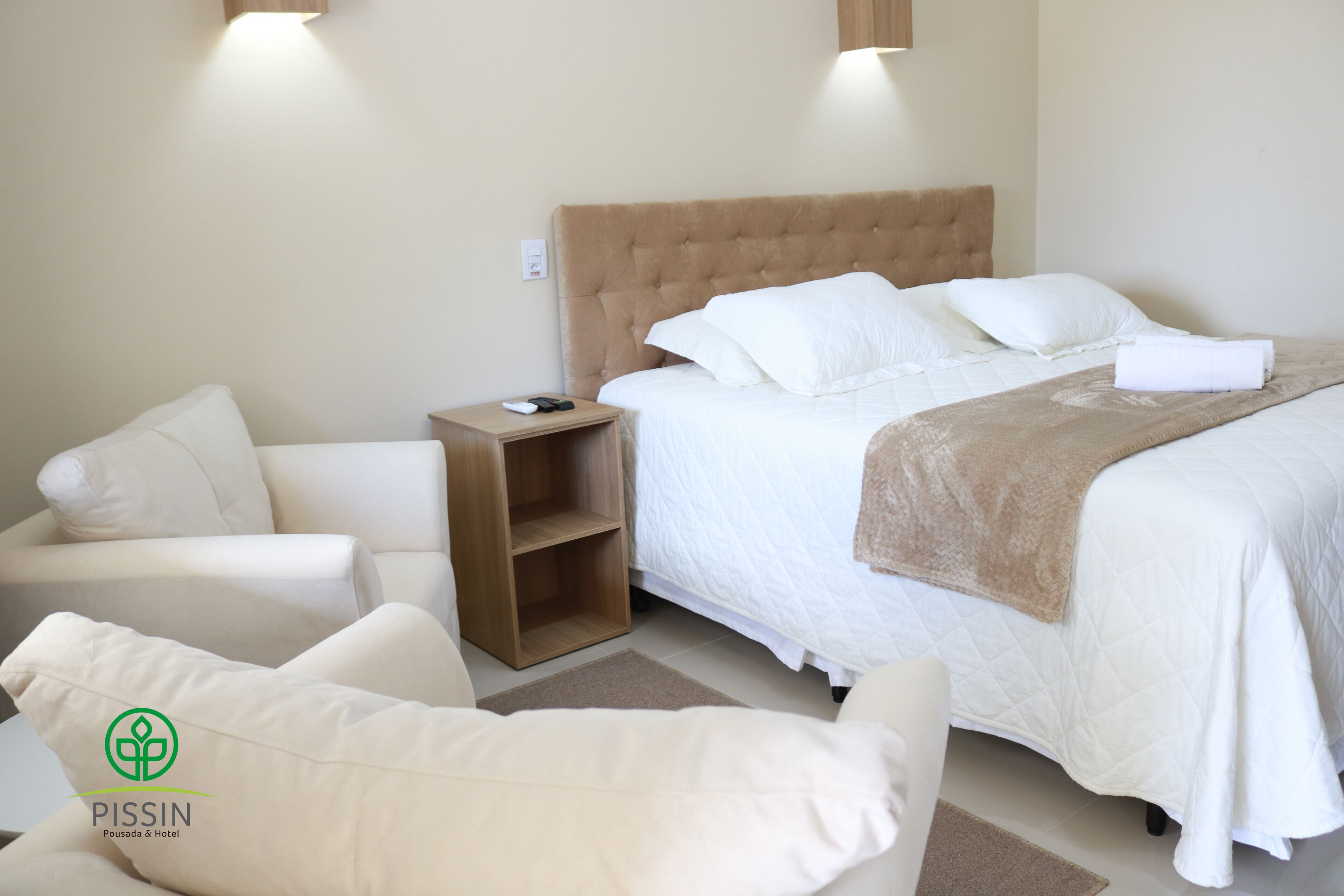 Una habitación de hotel con una cama, una mesita de noche de madera, un sillón blanco y un estante pequeño.