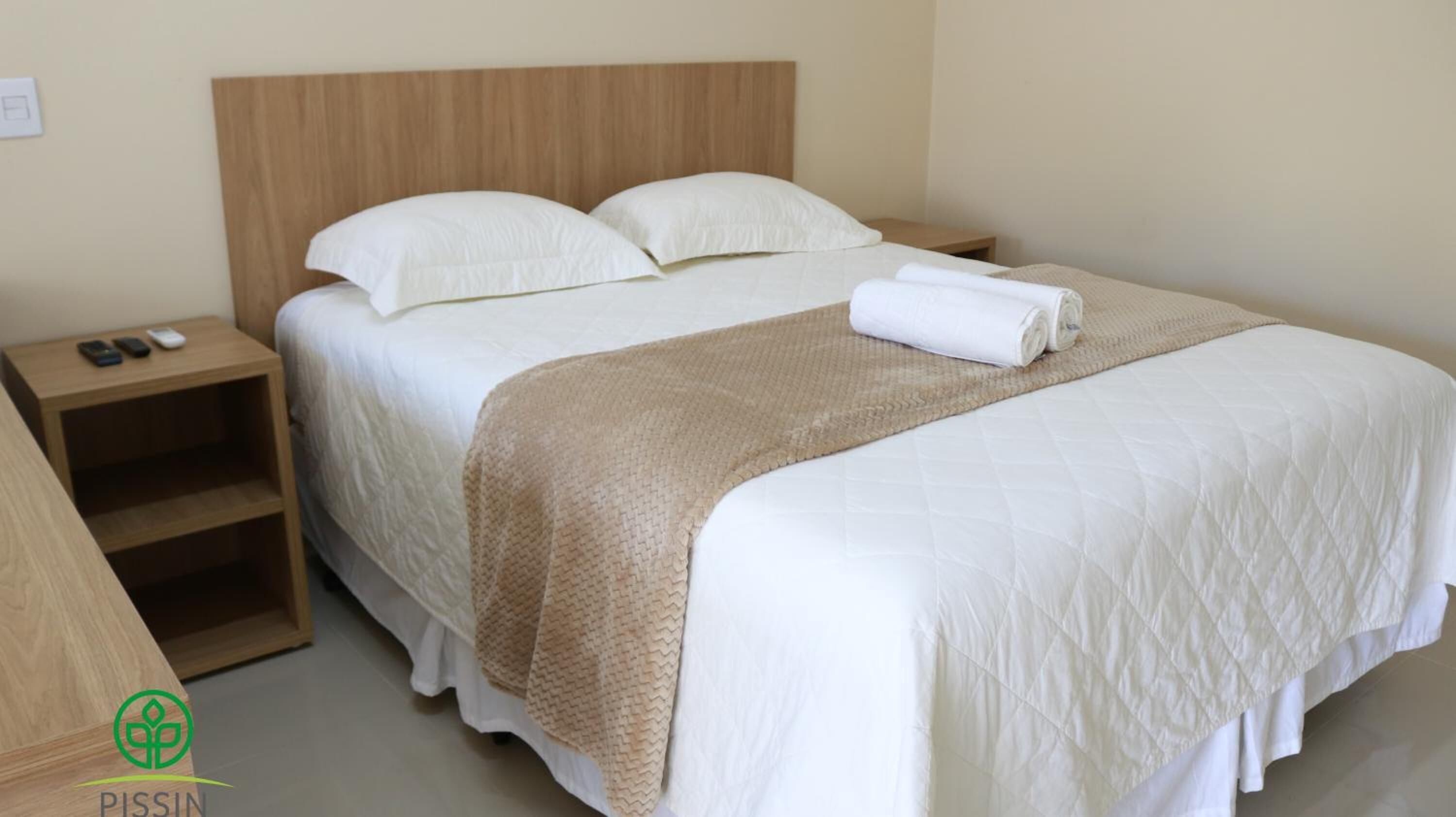 A neatly made bed with white linens and a beige throw blanket, a wooden nightstand with a remote control, and a small shelf unit.