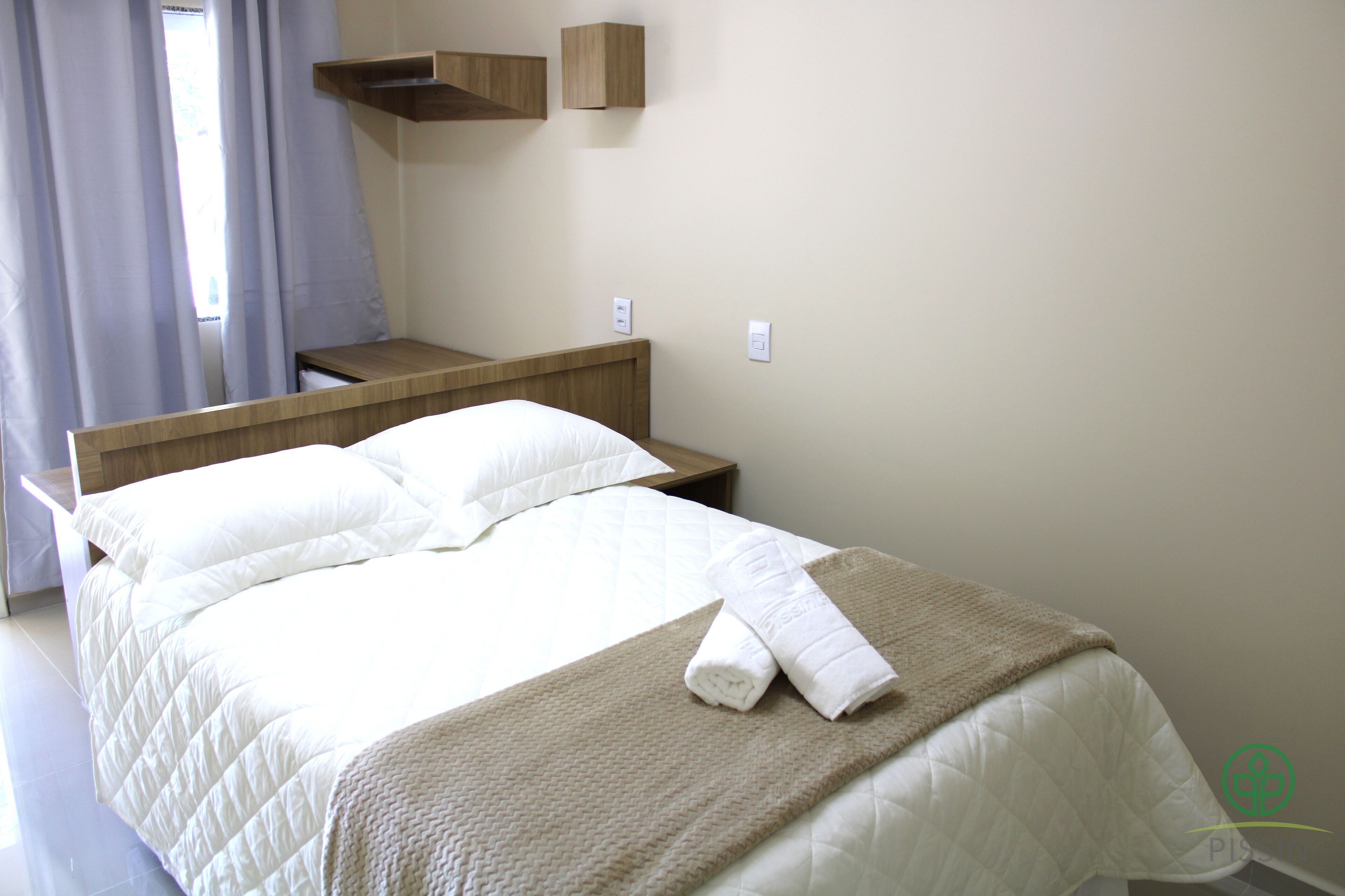 Una cama bien hecha con sábanas blancas y una manta color beige, una mesita de noche de madera y un estante fijado a la pared.