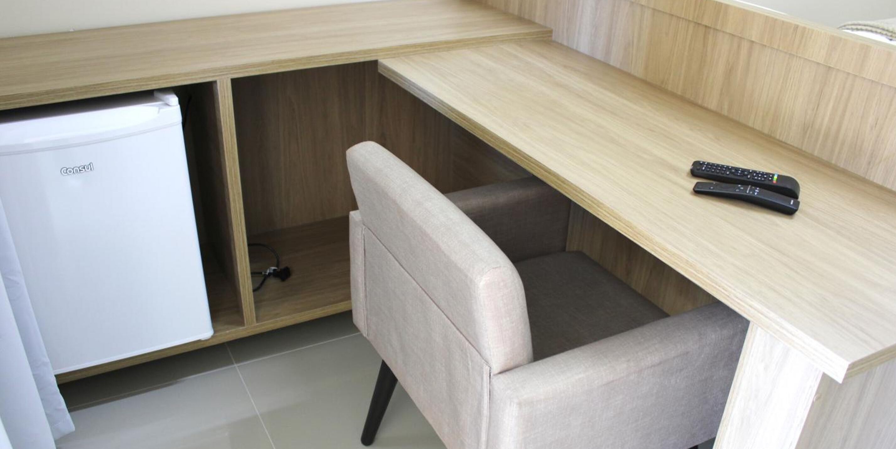 A compact kitchenette with a fridge, a chair, and a remote control on a wooden desk.