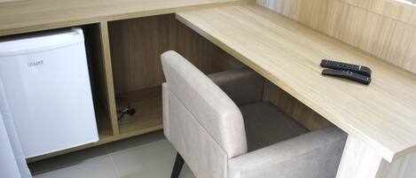 A compact kitchenette with a fridge, a chair, and a remote control on a wooden desk.