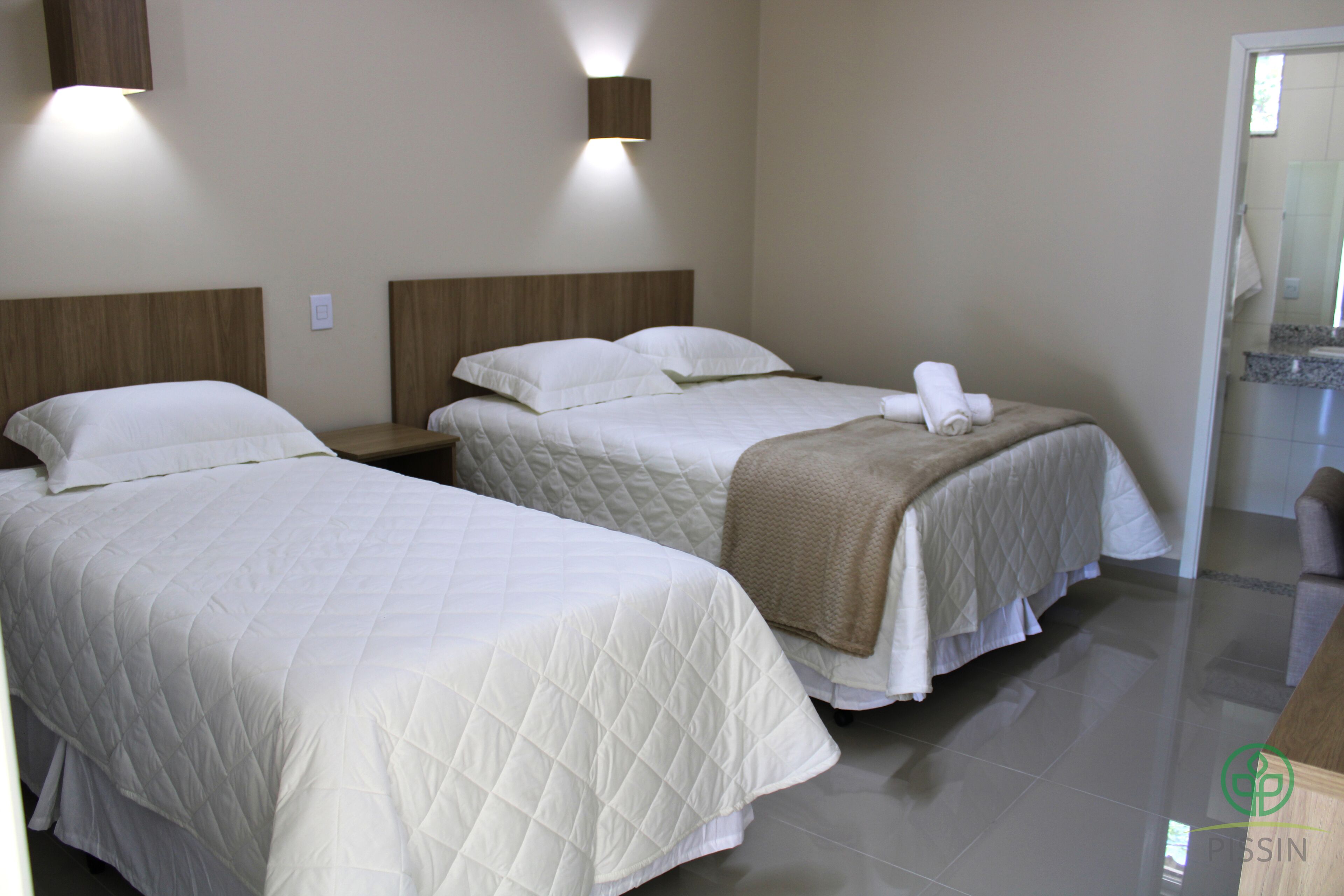 Habitación de hotel con dos camas, cada una con colcha blanca acolchada y manta marrón. Hay lámparas fijadas a la pared sobre cada cama.