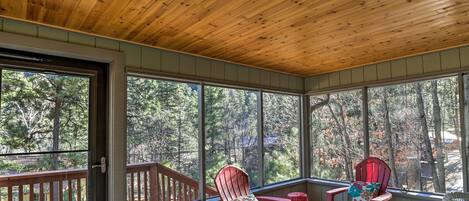 A covered porch with two red Adirondack chairs, a small table with pinecones, and a view of trees through large windows.