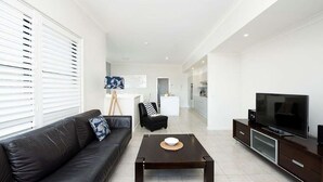 Un salon moderne avec un canapé en cuir noir, une table basse, un téléviseur et une cuisine en arrière-plan.