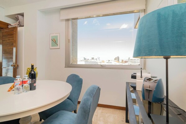 Quarto de hotel com uma mesa redonda, cadeiras azuis, uma janela com vista para a cidade, uma luminária e uma impressora.