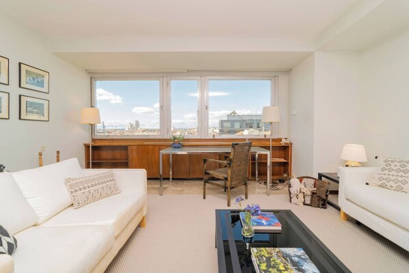 A modern living room with a sofa, a coffee table, a desk, and a chair. There are two windows with a view of the city.