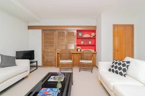 Een woonkamer met een bank, een salontafel, een televisie, twee rieten fauteuils, een rode plank met decoratieve objecten, en een houten deur.