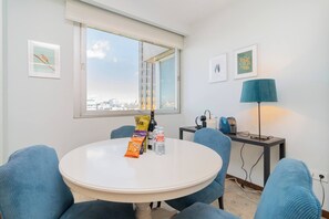 Una habitación con una mesa redonda, sillas, un ventanal con vista a la ciudad, un escritorio con lámpara y cuadros en la pared.