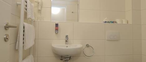 A bathroom with a sink, toilet, and a mirror. There are towels hanging on a rack, a trash bin, and a soap dispenser.