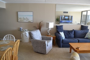 Ein Wohnzimmer mit einem blauen Sofa, einem grauen Sessel, einem Holztisch, einem Fernseher auf einem Lowboard und einem Gemälde an der Wand.
