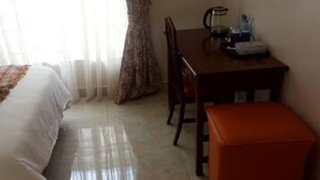 Habitación de hotel con cama, una mesa de madera con cafetera, una botella de agua y un pequeño reposapiés naranja.