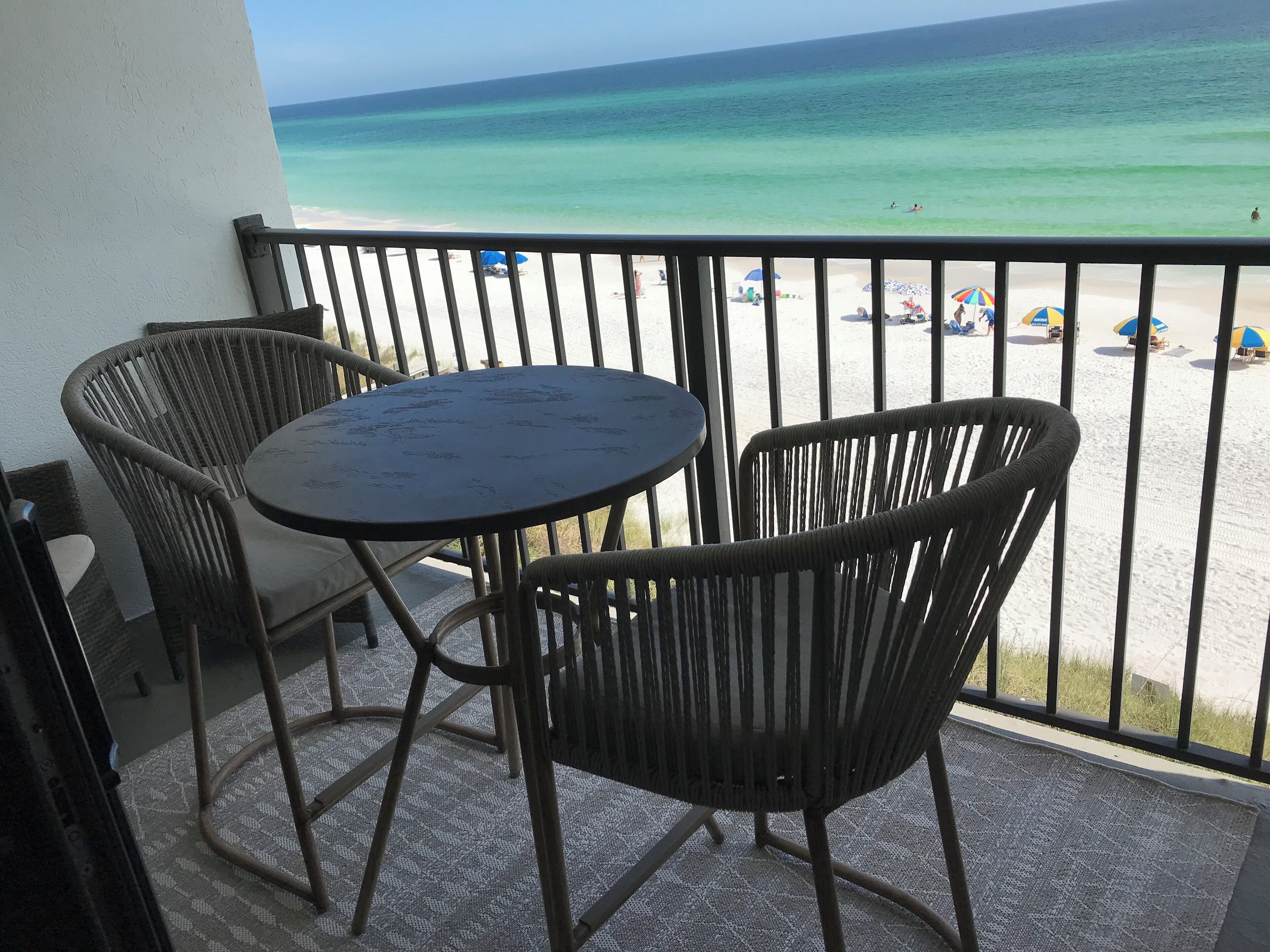 A balcony with two wicker chairs and a small round table, overlooking a beach with umbrellas and people.