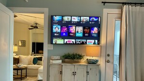 Una camera d'albergo con una televisione che mostra diversi canali in streaming, un letto, un comodino con una lampada, una piccola pianta e vista all'esterno attraverso una finestra.
