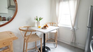 Una habitación pequeña y luminosa con una mesa blanca y dos sillas de madera, un florero con flores y una ventana con cortinas blancas.
