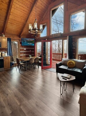 Una cabaña espaciosa con interior de madera, un comedor, un sofá y ventanas amplias que ofrecen vistas al exterior.