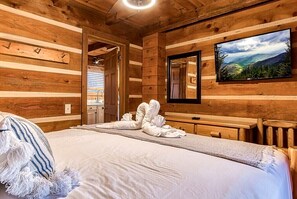 A wooden cabin room with a bed, towels, a television, and a window.