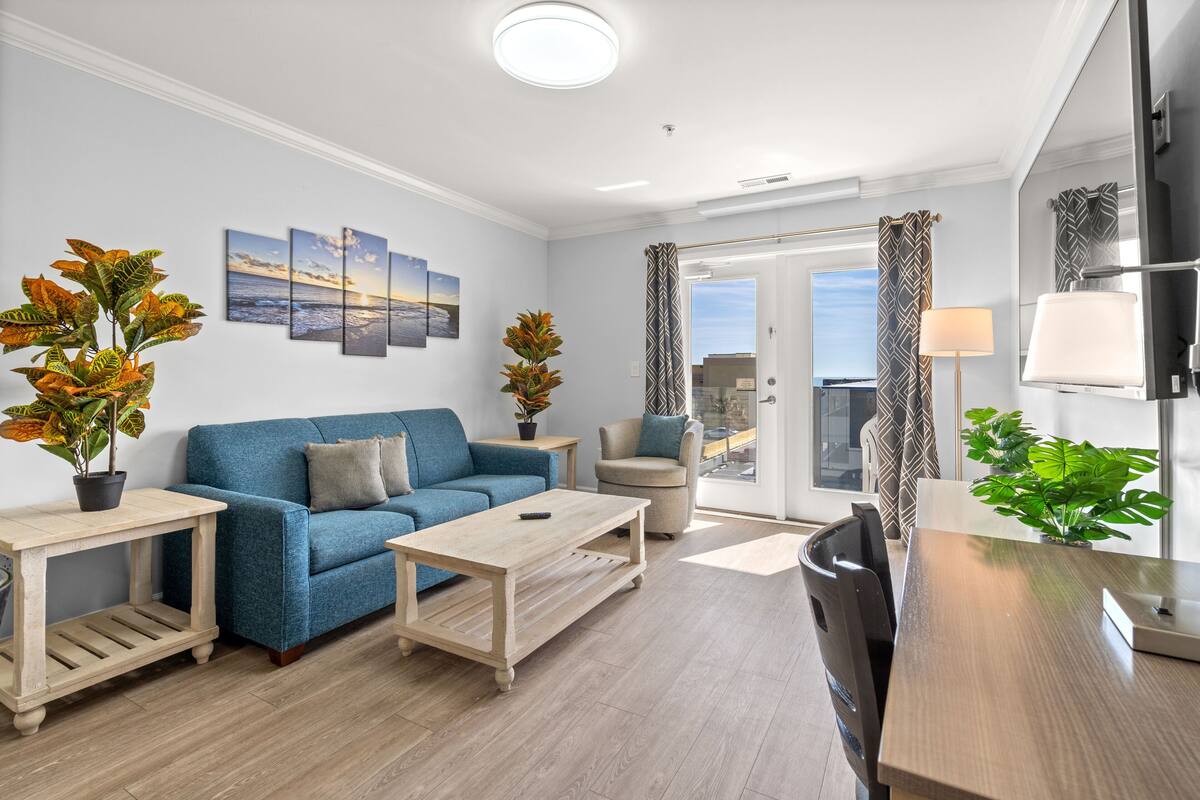 A modern living room with a blue sofa, wooden coffee table, and a dining area with a wooden table and chairs. There are potted plants and artwork on the walls.