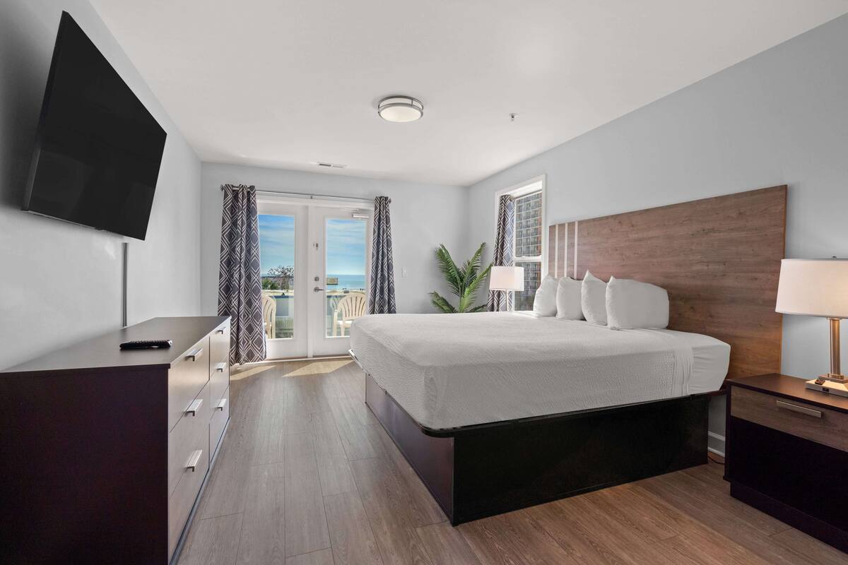 A modern hotel room with a large bed, a flat-screen TV, a wooden headboard, and a balcony with curtains.