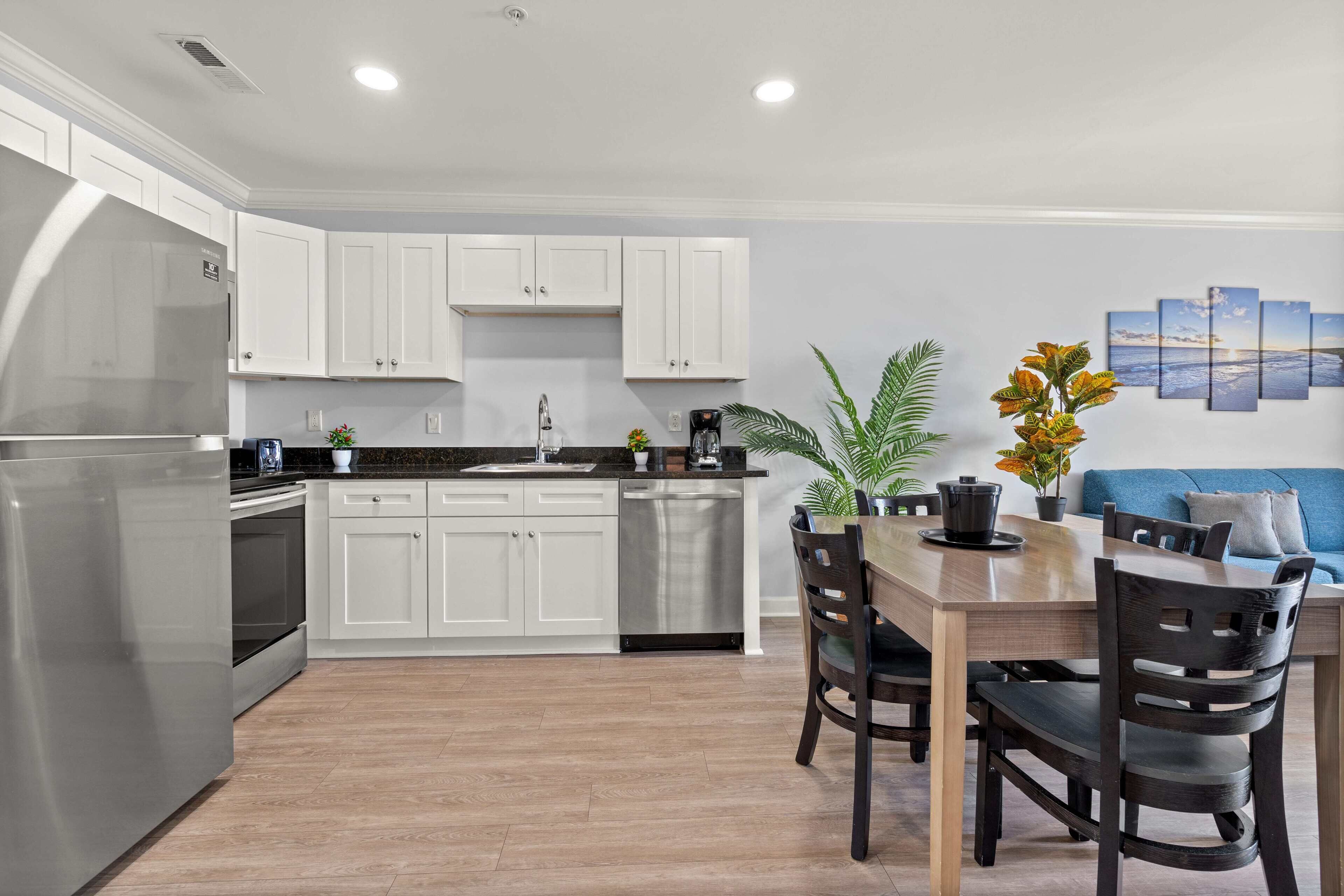 Una cocina moderna con armarios blancos, un refrigerador de acero inoxidable y un comedor con mesa y sillas de madera.