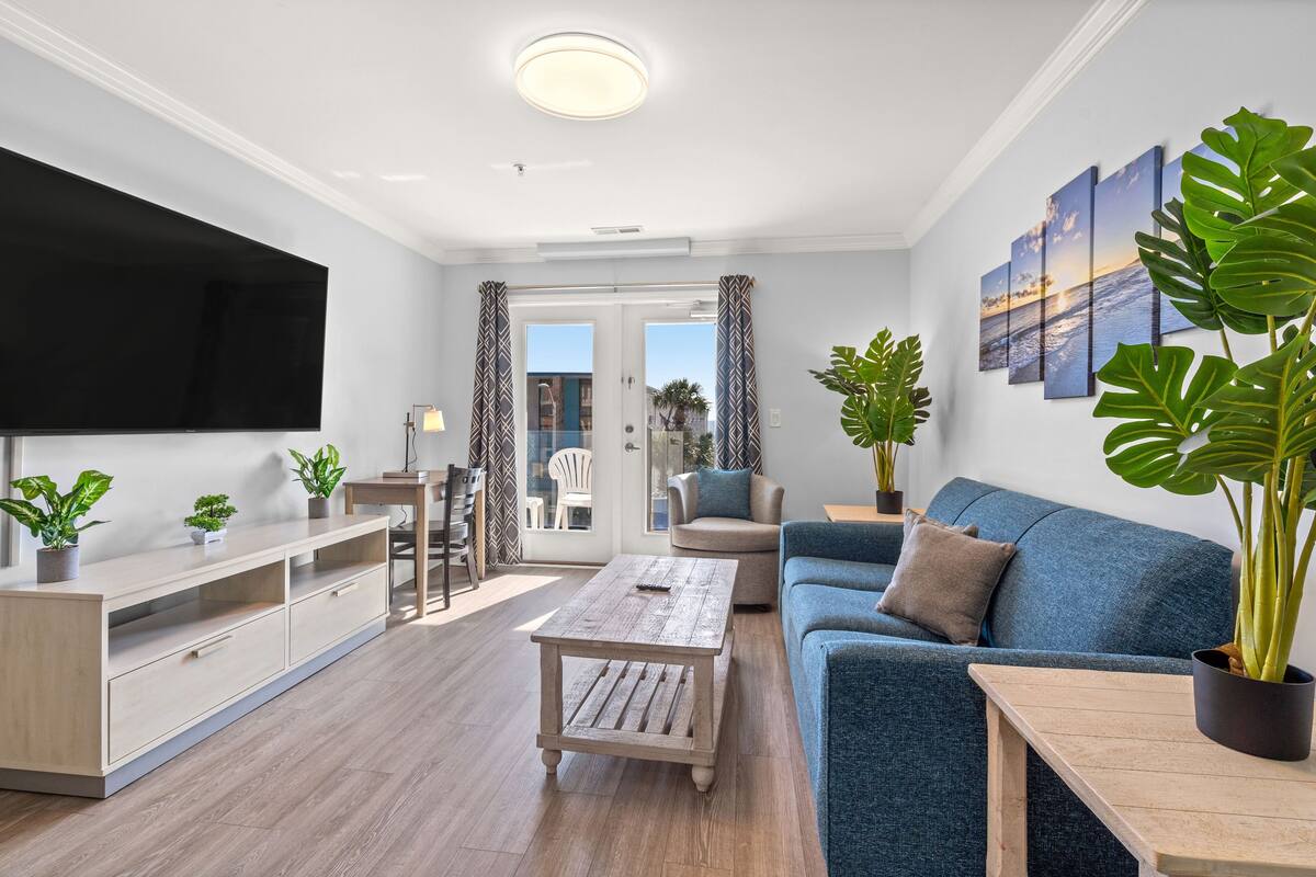 A modern living room with a flat-screen TV, a blue sofa, a wooden coffee table, and potted plants.
