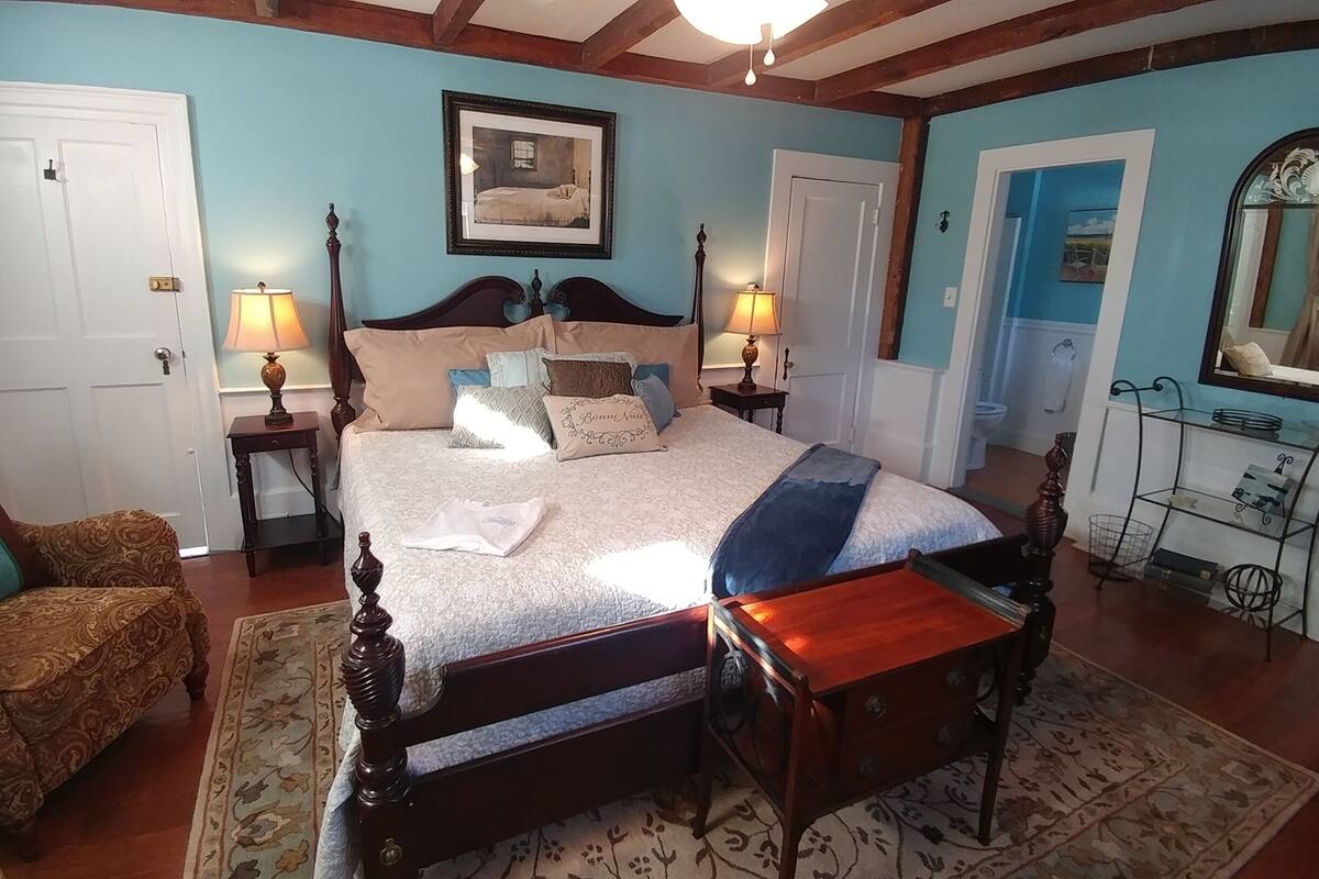 A bedroom with a large bed, two bedside tables, a chair, a wooden bench, a mirror, and a small shelf.