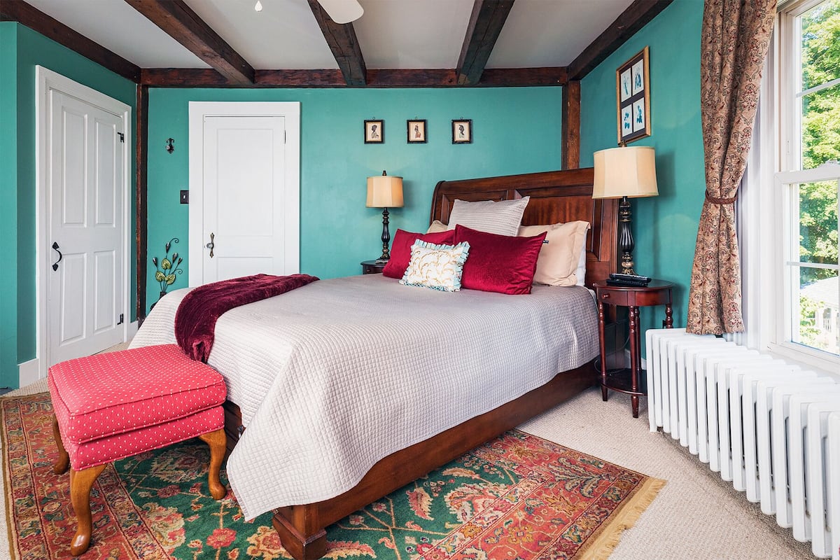 A bedroom with a large bed, two bedside tables, a red upholstered bench, a patterned rug, and a window with curtains.