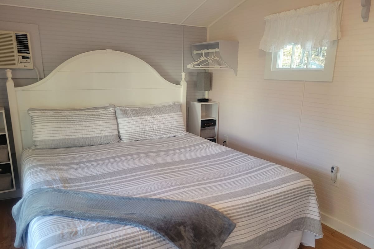 A neatly made bed with striped bedding, a bedside table with a lamp, a hanging clothes rack, and a window with a white valance.