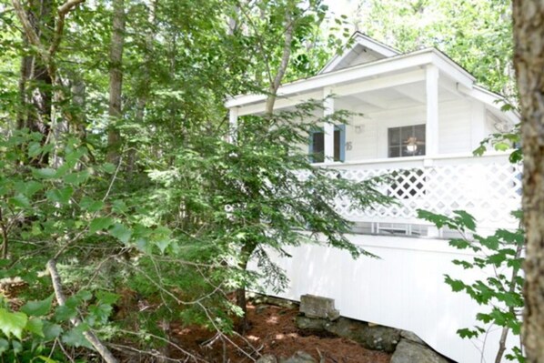 A white wooden cabin with a covered porch, surrounded by dense forest. - 2 River Road Inn and Cottages (Cape Neddick)