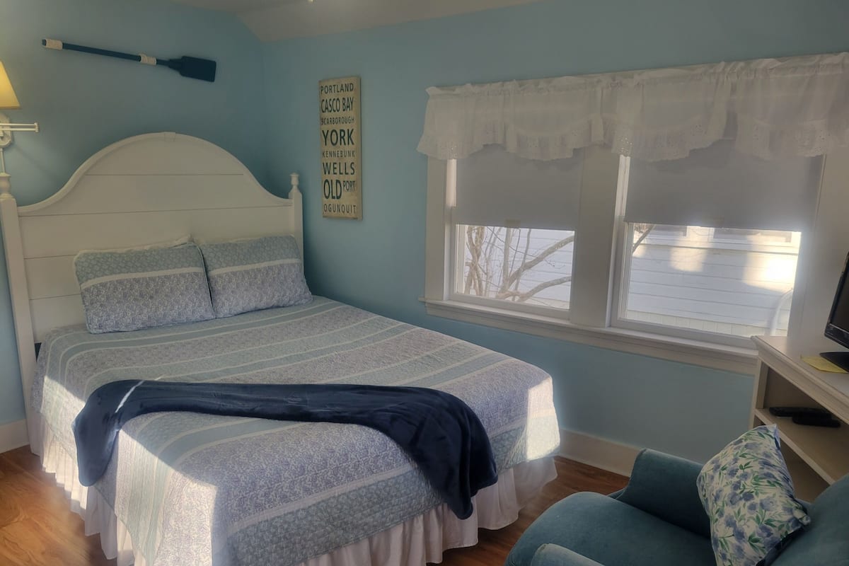A bedroom with a bed, a window with a view, a blue armchair, and a wall sign listing city names.