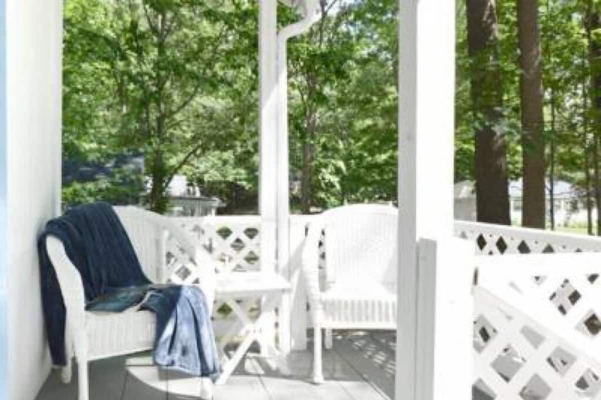 A white wooden porch with a bench and a table, surrounded by a lattice fence, set in a wooded area.