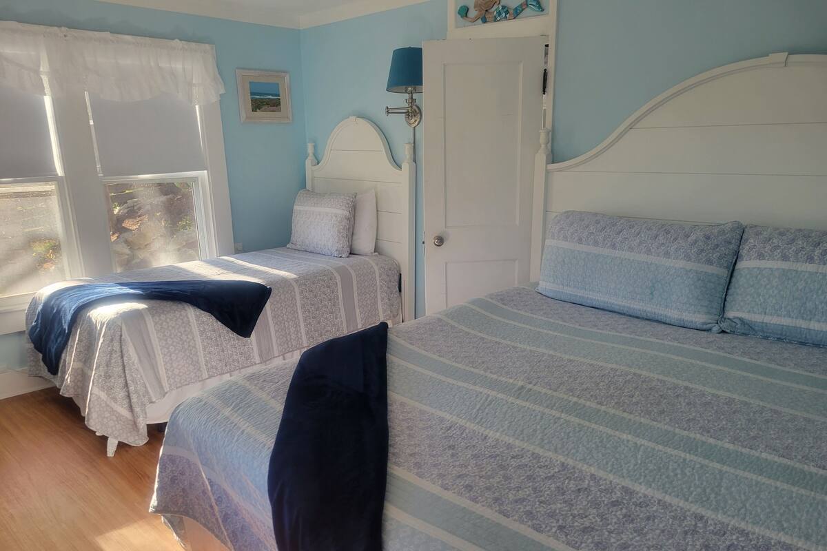 A bedroom with two beds, a window with curtains, a door, and a wall-mounted lamp.