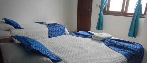 Habitación de hotel con dos camas, cada una con ropa de cama blanca y azul, una puerta de madera y un ventanal con cortinas.