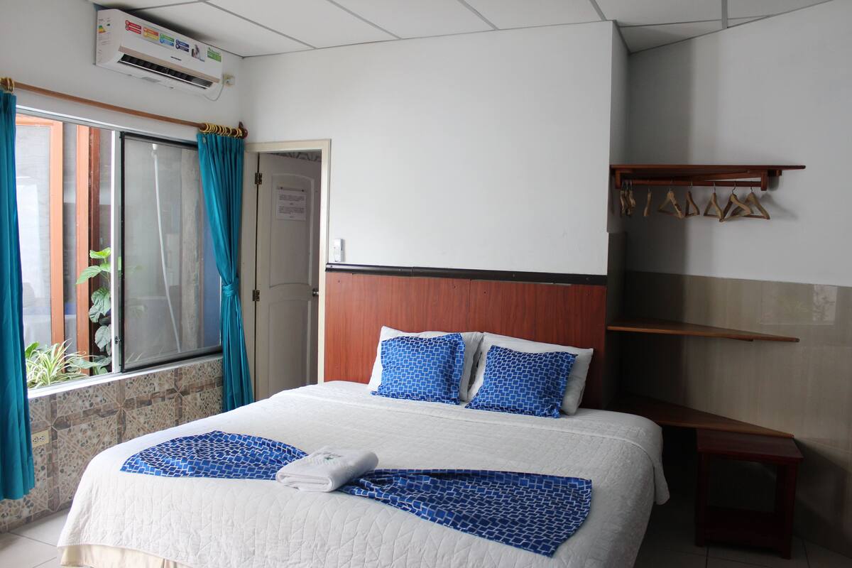 A hotel room with a bed, a wooden headboard, a window with curtains, a small table, and a wall-mounted shelf.