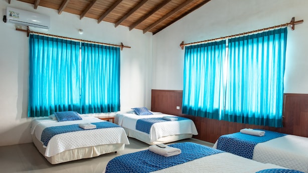 A hotel room with two beds, blue curtains, wooden ceiling beams, and a white air conditioning unit. - Hotel La Gran Tortuga (Puerto Villamil)