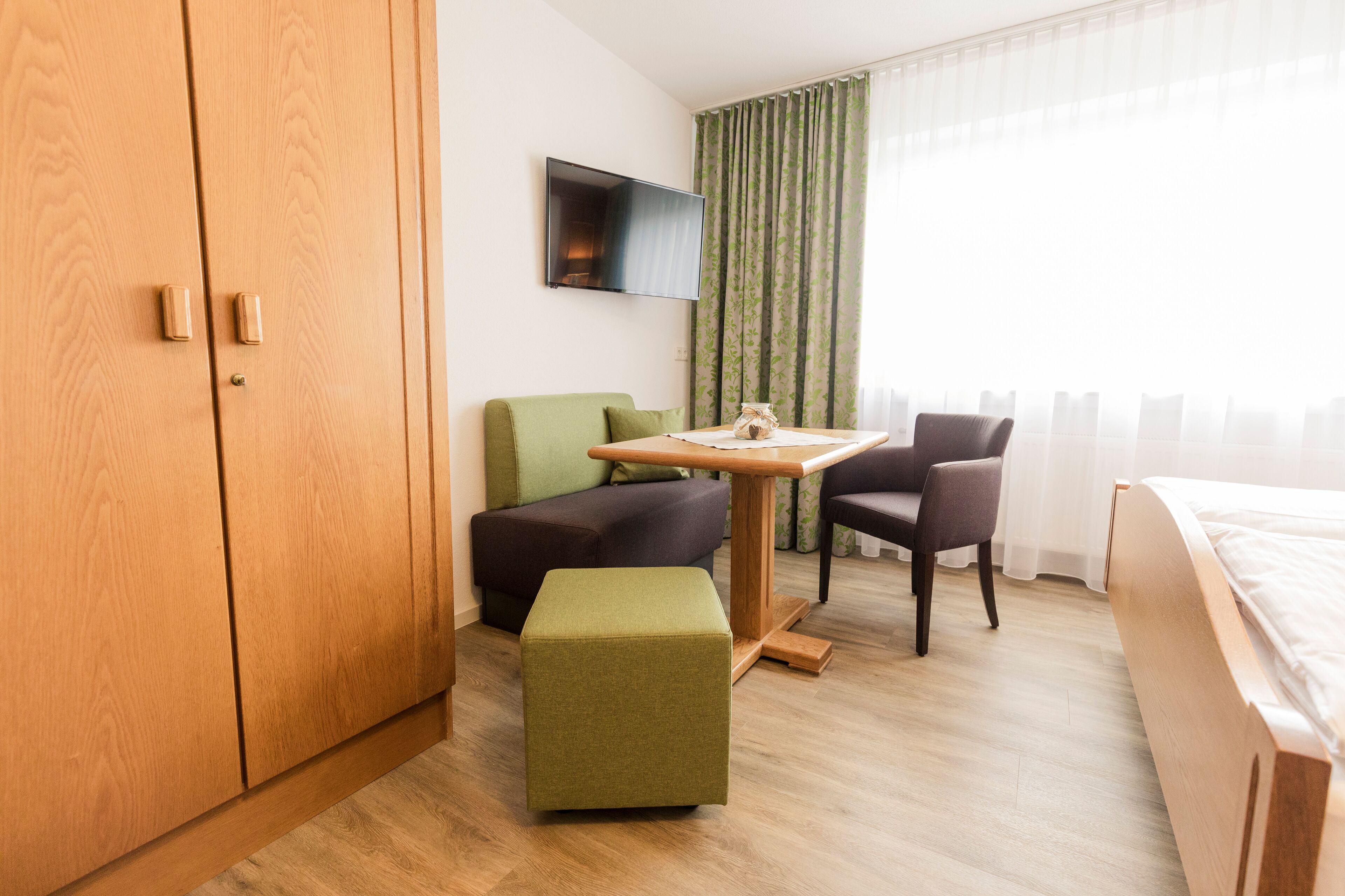 Ein Hotelzimmer mit Holzschrank, grünem Ottomanen, Holztisch mit Lampe, Wand-Fernseher und Bett mit weißer Bettwäsche.
