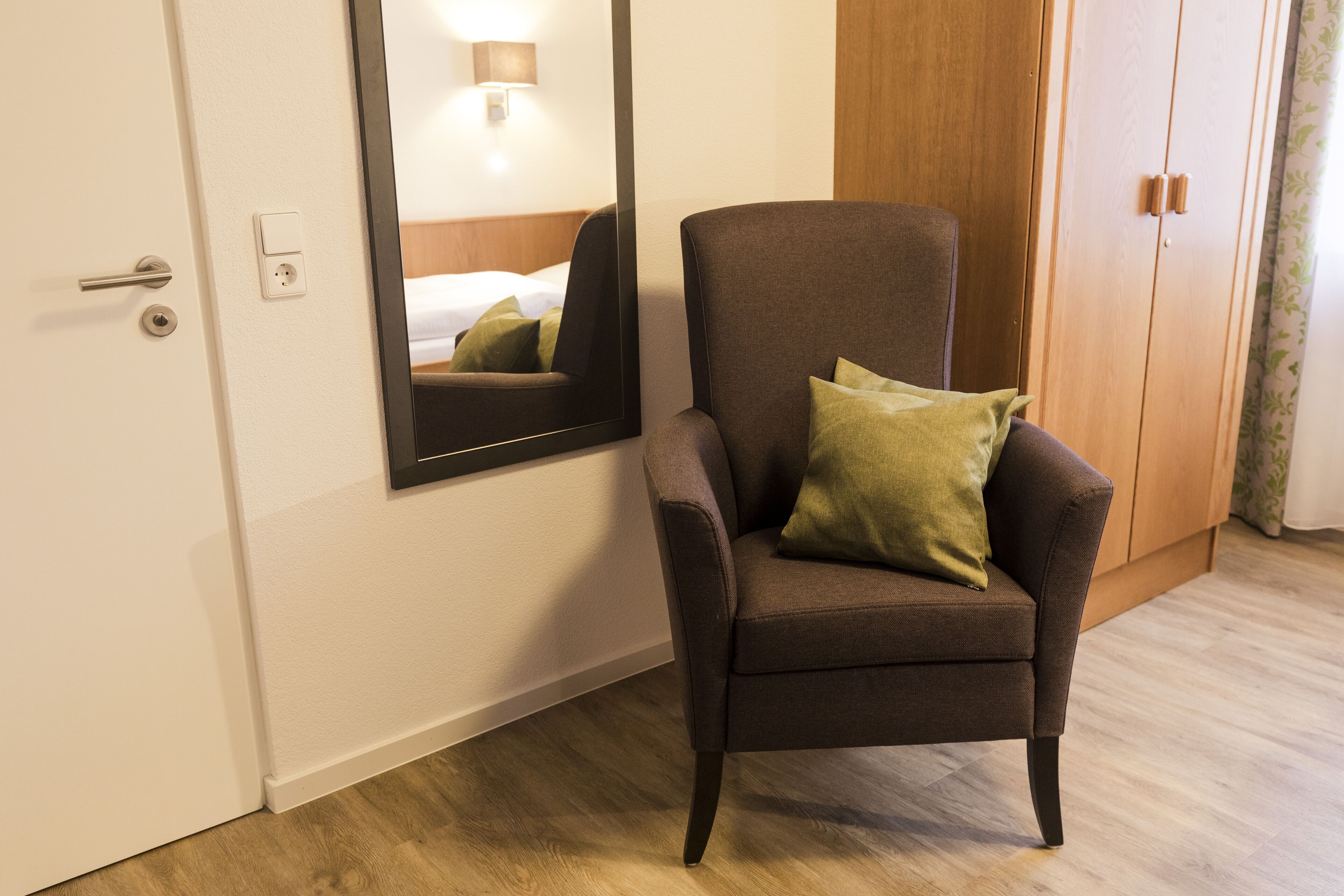 Una habitación de hotel con un sillón marrón, un espejo, un armario de madera y una puerta blanca.