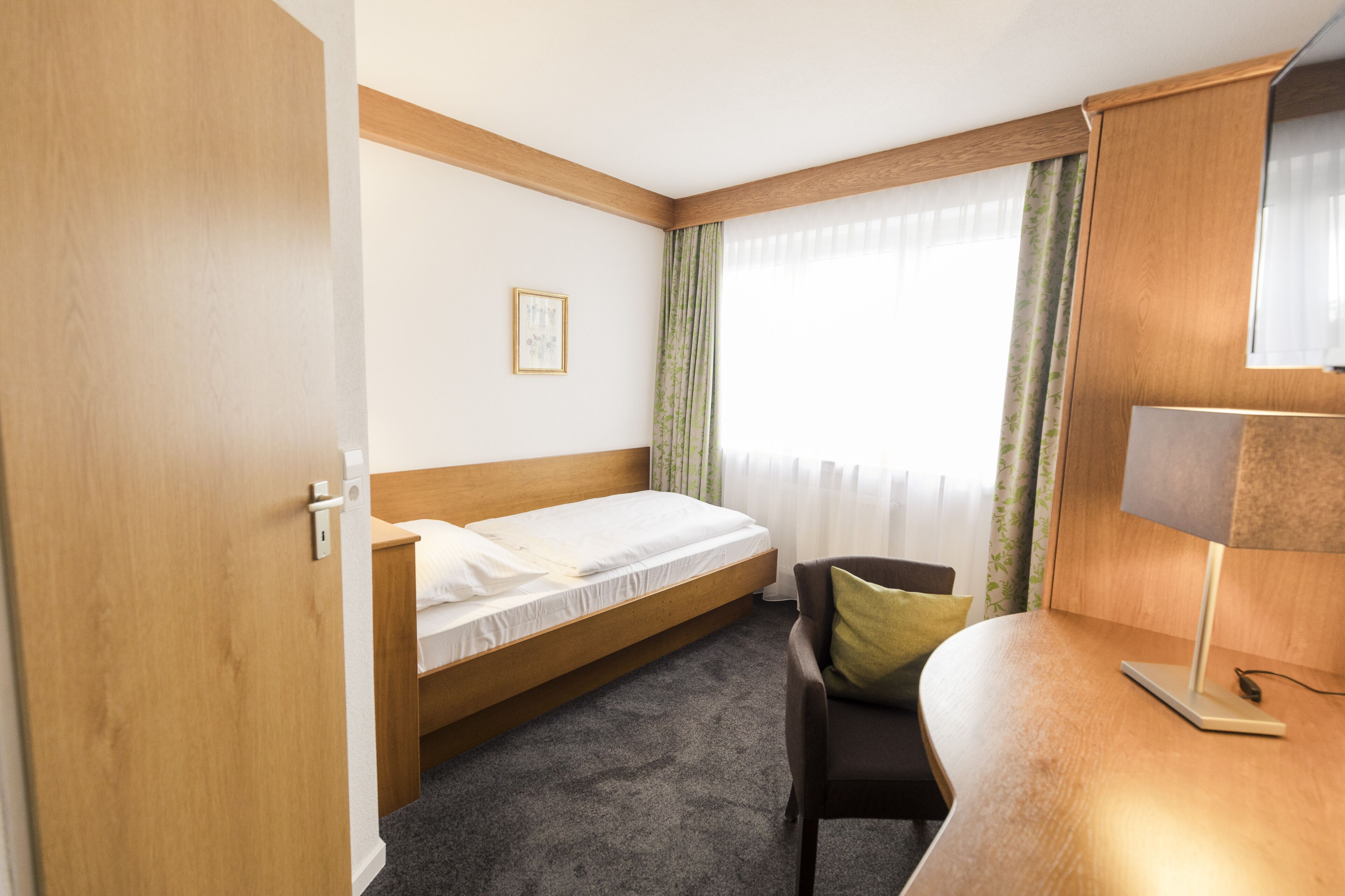 A hotel room with a single bed, a desk with a lamp, a chair, and a window with curtains.