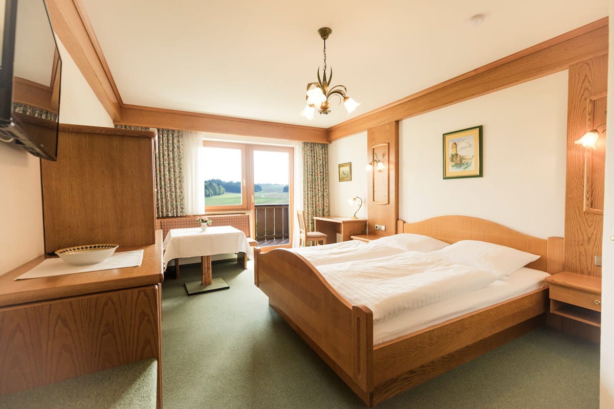 A hotel room with a wooden bed, a desk, a chair, a small table, a window with a view, and a chandelier.