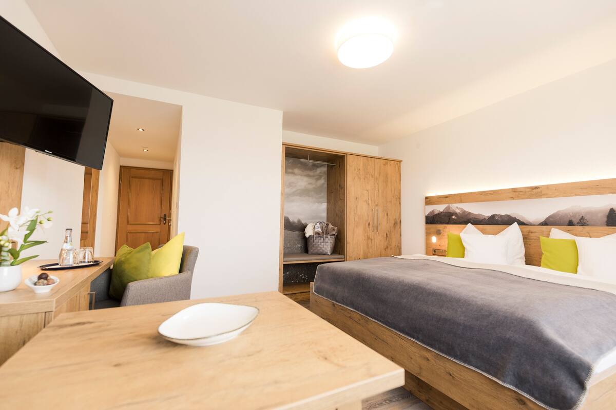 A modern hotel room with a bed, a TV, a wooden desk, and a view of mountains.