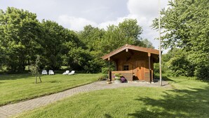 Uma cabana de madeira com telhado inclinado, cercada por um gramado bem cuidado e árvores.
