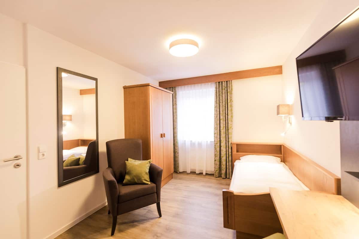 A hotel room with a single bed, a chair, a desk, a wardrobe, a mirror, and a window with curtains.