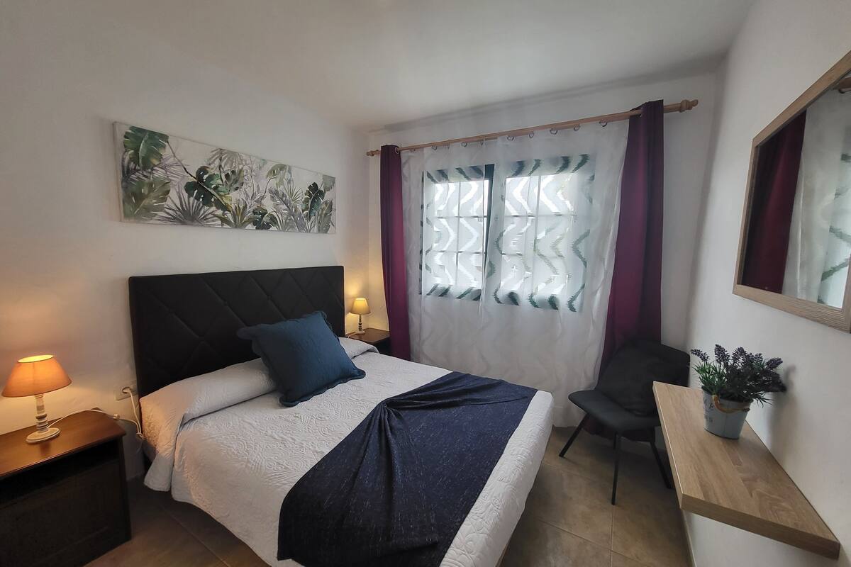 A neatly arranged bedroom with a bed, bedside lamp, a small table with a plant, and a window with curtains.
