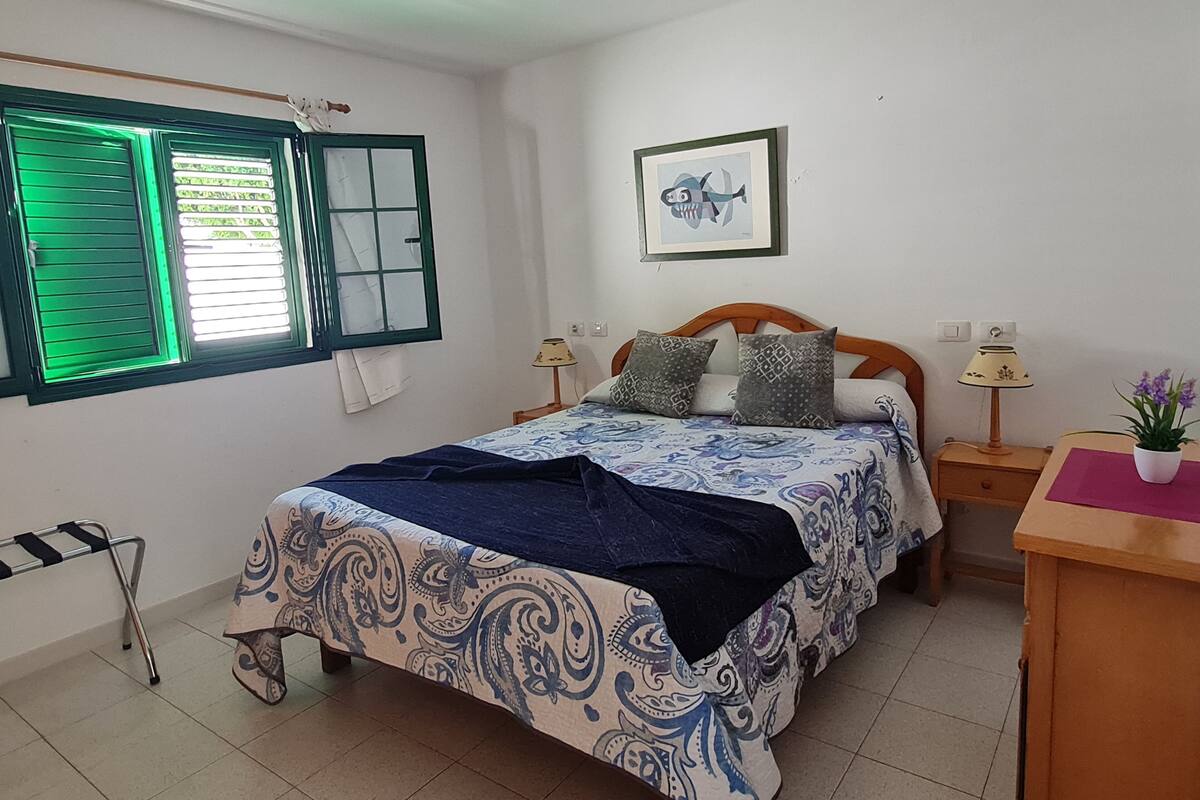 A bedroom with a bed, two bedside tables, a window with green shutters, a framed picture on the wall, and a potted plant on the nightstand.