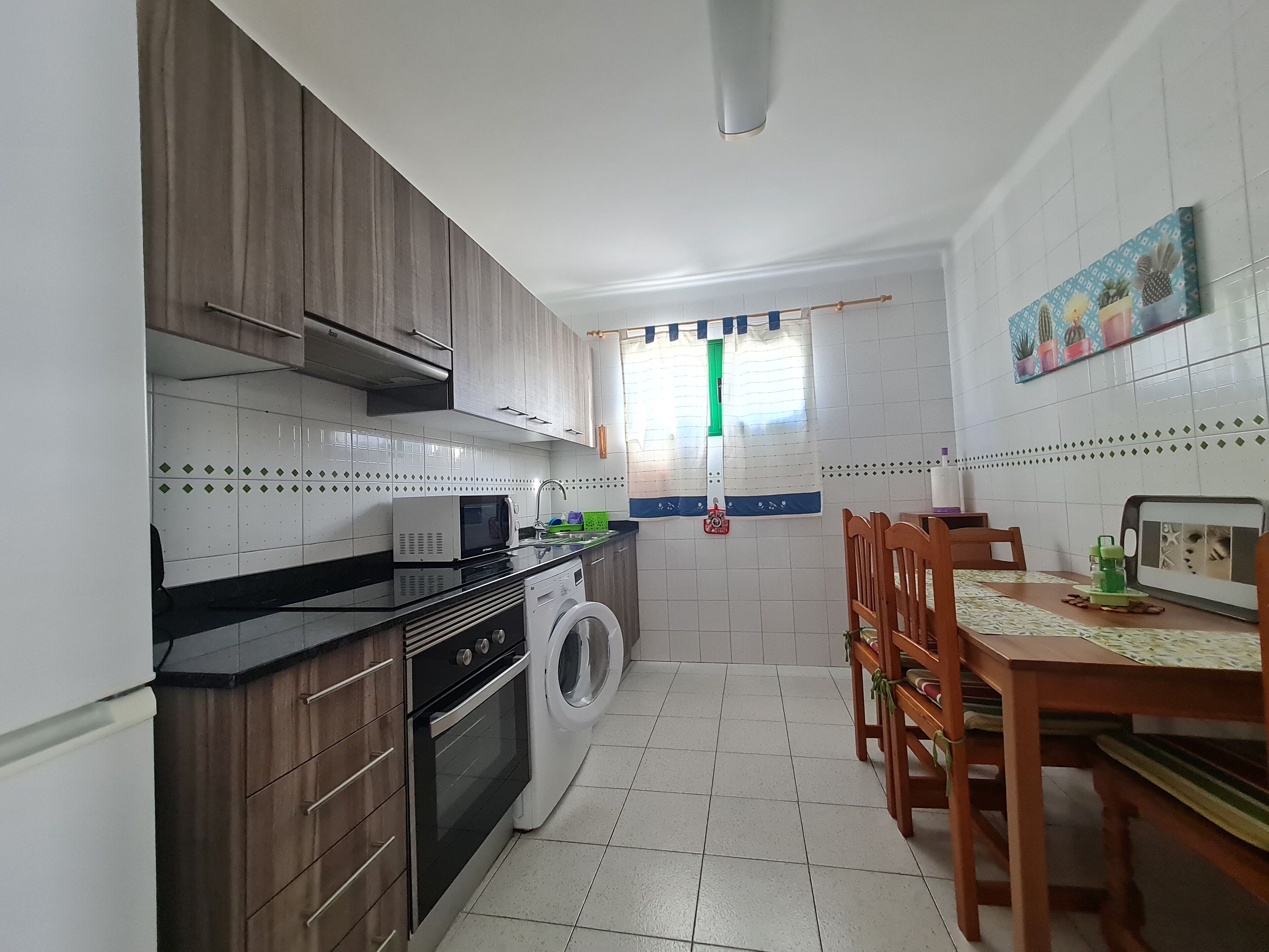 A compact kitchen with a washing machine, a microwave, and a stove. There is a dining table with chairs and a window with curtains.
