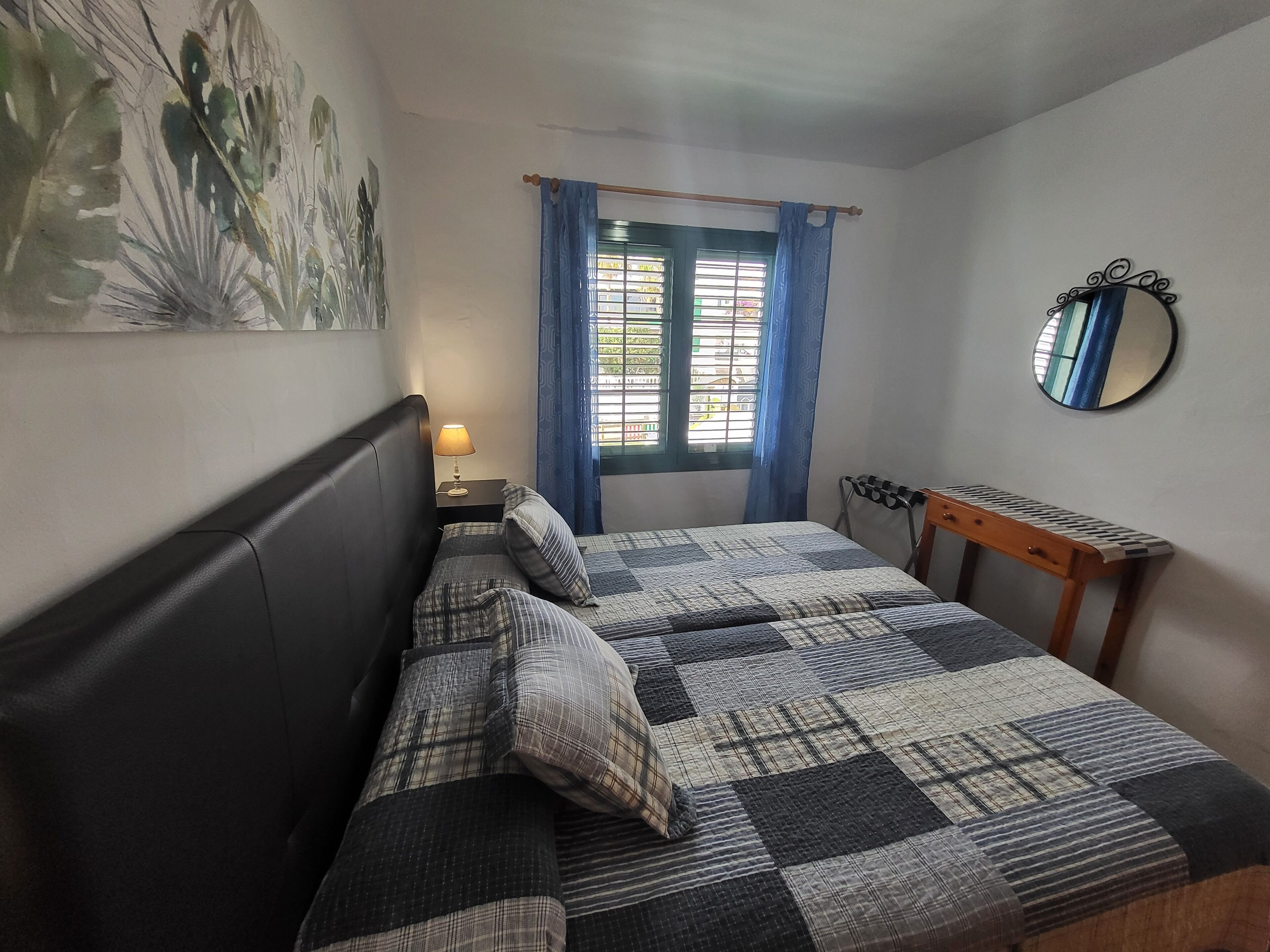 A bedroom with a bed, a nightstand with a lamp, a window with curtains, a mirror, and a wall-mounted shelf.