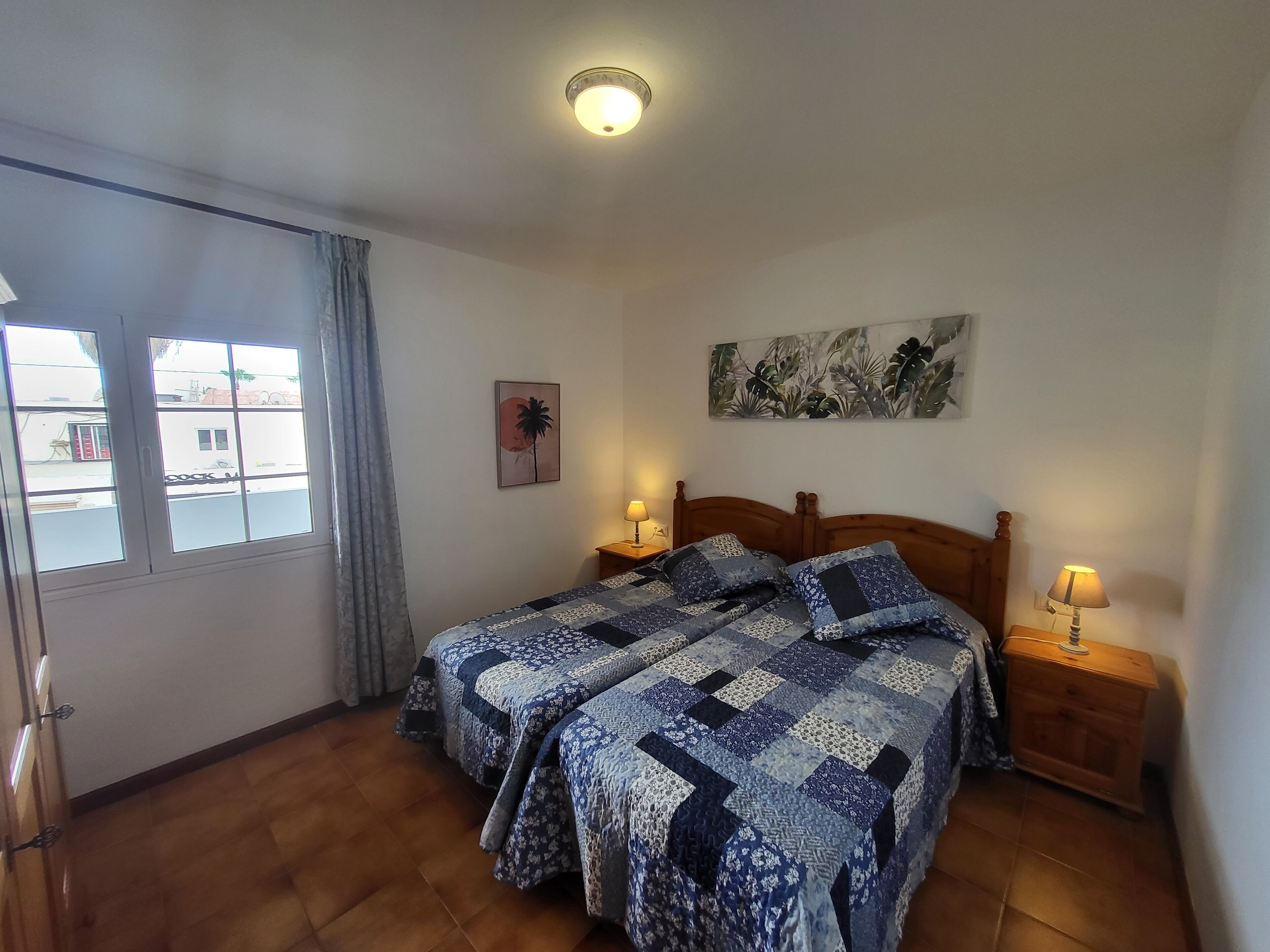 Une chambre avec deux lits, une fenêtre avec des rideaux, un luminaire au plafond et une plante fixée au mur.
