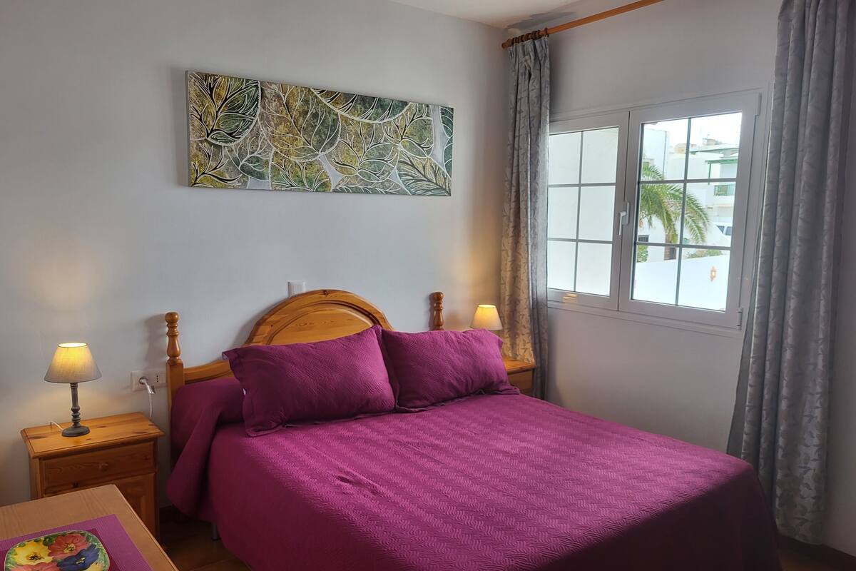 A bedroom with a bed, two bedside lamps, a nightstand, a table with a decorative plate, a window with curtains, and a wall art piece.