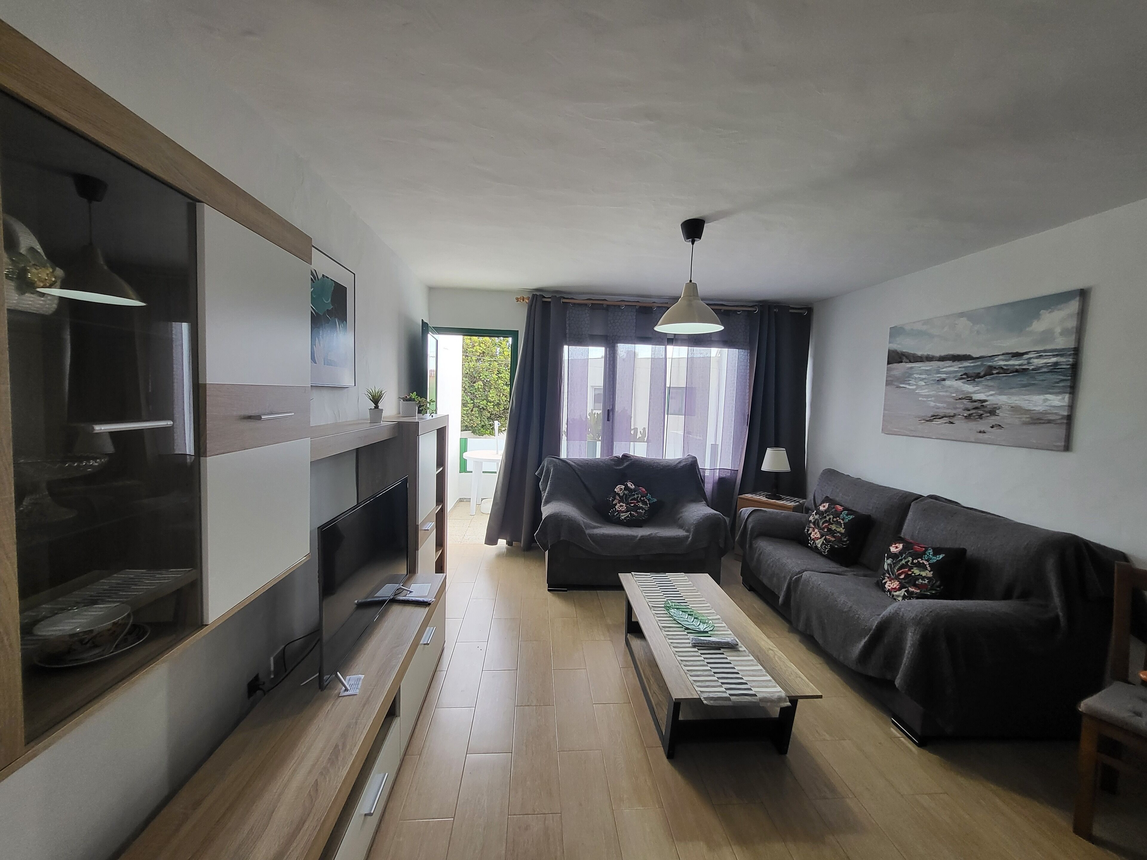 A living room with a sofa, a coffee table, and a TV. There is a window with curtains and a painting on the wall.