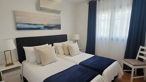 Habitación de hotel con dos camas, una mesita de noche con lámpara, una silla, un cuadro en la pared y una ventana con cortinas.